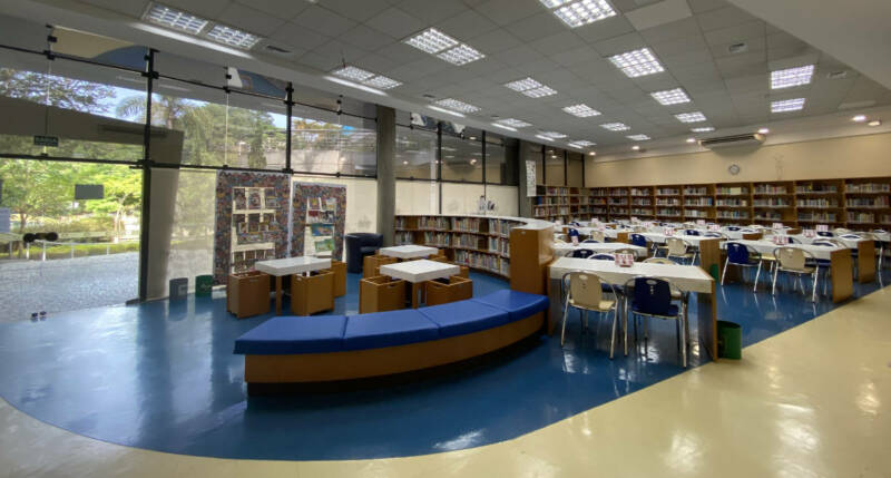 Biblioteca do Clube Paineiras - A imagem mostra uma área com um sofá à frente, ao centro estão mesas e cadeiras e ao fundo uma prateleira repleta de livros.