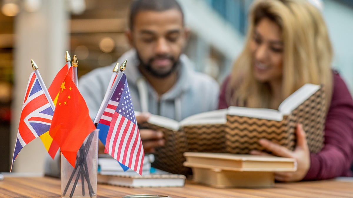 Foto de pequenas bandeiras de diversos países apoiados denteo de um copo, e ao fundo um casal estudando