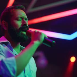 Homem cantando em palco com microfone, sob iluminação colorida em tons de neon, durante apresentação musical em ambiente fechado.