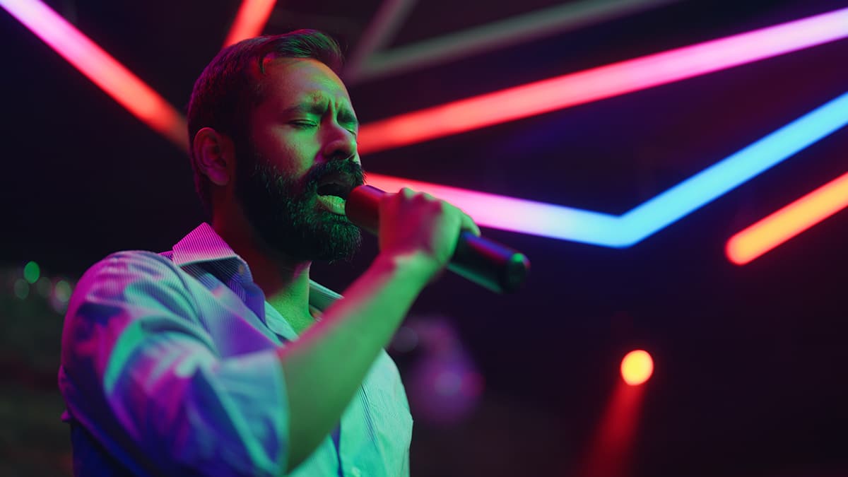 Homem cantando em palco com microfone, sob iluminação colorida em tons de neon, durante apresentação musical em ambiente fechado.