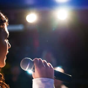 Mulher cantando ao microfone em palco iluminado, vista de perfil, com luzes fortes ao fundo e clima de apresentação ao vivo.
