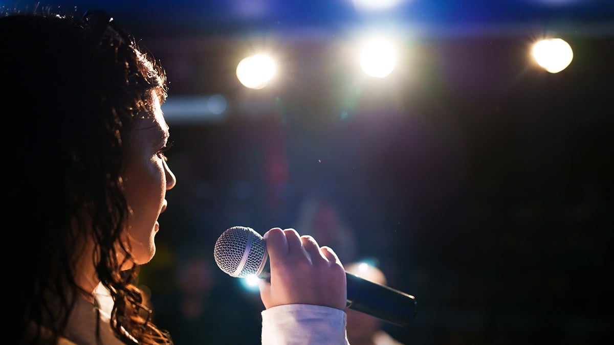 Mulher cantando ao microfone em palco iluminado, vista de perfil, com luzes fortes ao fundo e clima de apresentação ao vivo.